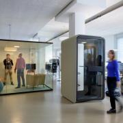 Unser Open Space Arbeitsplatz am Open Systems Standort in Bern. Ein Großraumbüro mit weißen Wänden, beigebraunem Boden, blauen Stühlen und weißen Arbeitstischen. Mittig darin ein Glaskubus mit weißer Rückwand als Besprechungsraum, der gerade von zwei Kollegen genutzt wird. Etwas rechts davon ein weiterer kleinerer Kubus, der als geräuschfreier Telefonraum dient.